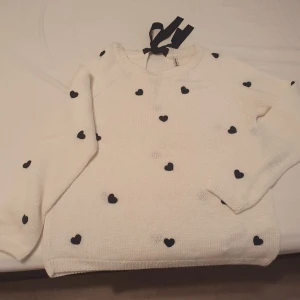 Stradivarius  - Superfin vit stickad tröja med små svarta hjärtan över hela. Perfekt för en mysig dag hemma eller en fika med vänner. Tröjan har långa ärmar och en söt rosett i nacken. Den är både bekväm och stilren, lätt att matcha med jeans eller kjol.