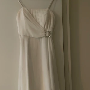 Vit klänning med smala axelband - Elegant vit klänning med smala axelband och v-ringad design. Den har en vacker detalj med en glittrig spänne framtill och en knytning bak. Perfekt för sommarens fester eller studentfiranden. Klänningen är i mycket bra skick och har en luftig passform.