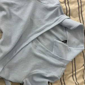 Ljusblå ribbad omlottopp - Säljer en superfin ljusblå ribbad omlottopp. Den har långa ärmar och en snygg omlottdesign som knyts i sidan. Perfekt för både vardag och fest! Toppen är i ett mjukt och stretchigt material som sitter bekvämt.