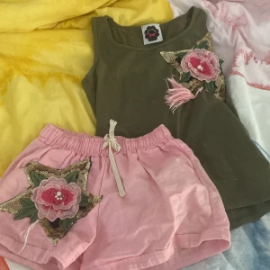 Grön linnetopp och rosa shorts med blommig detalj - Säljer ett set med en grön linnetopp och rosa shorts. Båda plaggen har en stor blommig applikation med paljetter och broderi. Toppen är ärmlös och shortsen har en bekväm resårmidja med knytband. Perfekt för sommardagar! Storlek xs men passar även s! Den är använd och man ser det lite på den, men inga skador! 