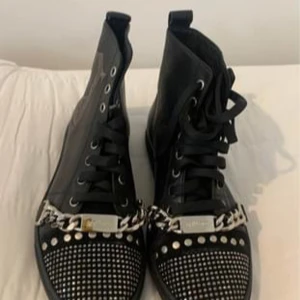 Svarta boots med kedjedetalj från Galliano - Snygga svarta boots i skinn från Galliano med coola kedjedetaljer och glittriga nitar på tån. De har snörning framtill och en rad med nitar längs baksidan. Perfekta för att ge din outfit en edgy touch! Storlek 38 dam  Inköpta i Tyskland