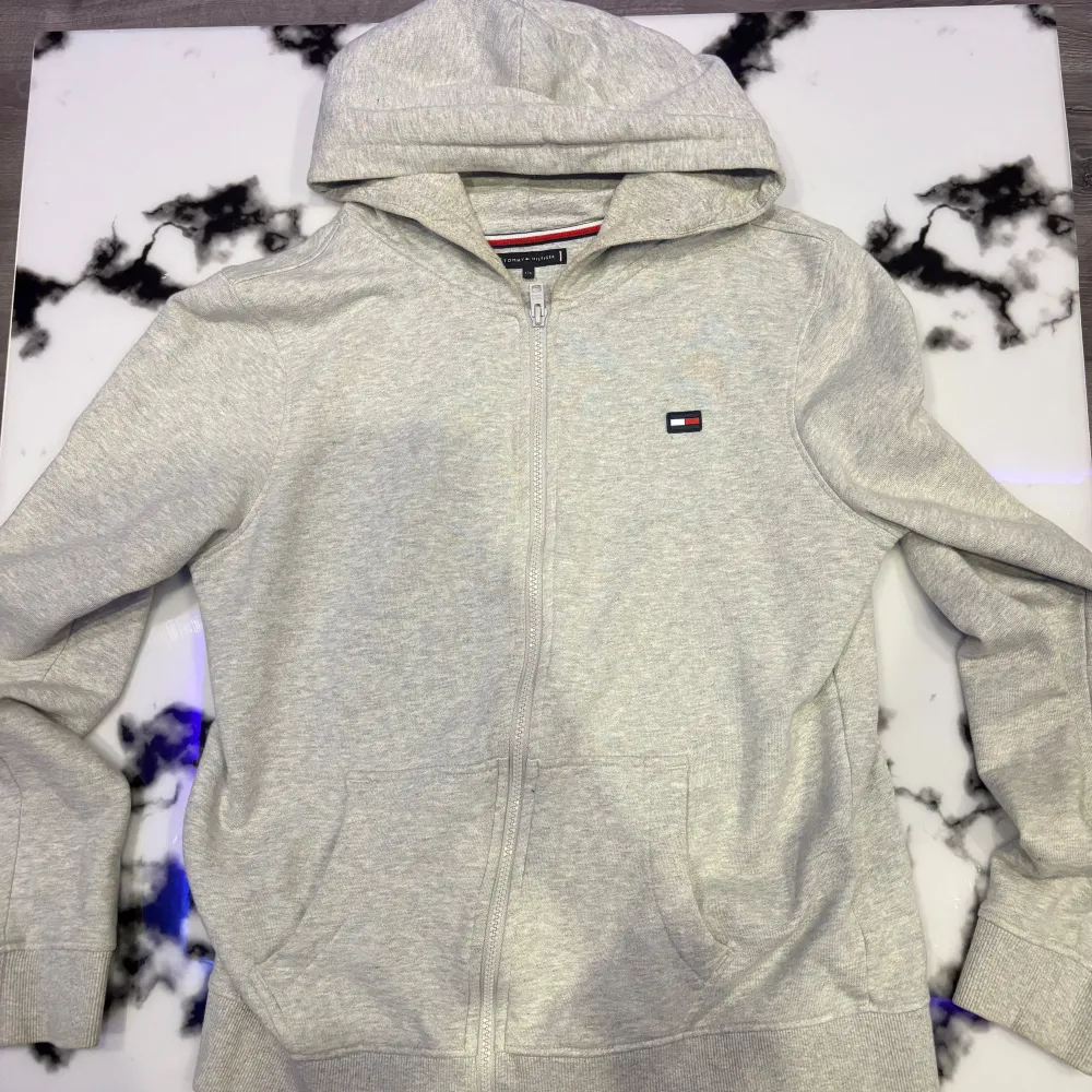 Säljer en grå hoodie från Tommy Hilfiger i mycket bra skick. Den har en dragkedja framtill och en klassisk logga på bröstet samt en stor logga på ryggen. Perfekt för både höst och vår. Jag bär normalt storlek S och den passar. Inte använd på snart ett år.. Tröjor & Koftor.