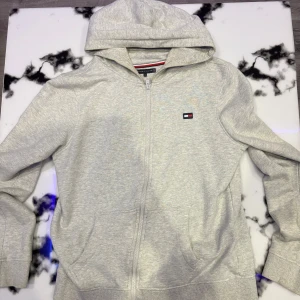 Tommy Hilfiger hoodie - Säljer en grå hoodie från Tommy Hilfiger i mycket bra skick. Den har en dragkedja framtill och en klassisk logga på bröstet samt en stor logga på ryggen. Perfekt för både höst och vår. Jag bär normalt storlek S och den passar. Inte använd på snart ett år.