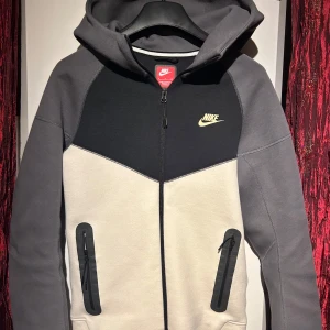 Nike tech fleece - Säljer en snygg och bekväm hoodie från Nike i grått och svart. Den har en dragkedja framtill och en klassisk Nike-logga i gult på bröstet. Perfekt för höst och vår, med långa ärmar och en skön huva. Passar både till träning och vardagsbruk. Storleken är inte angiven, men den ser ut att vara normal i passformen. Hör av er vid minsta fundering 🍾