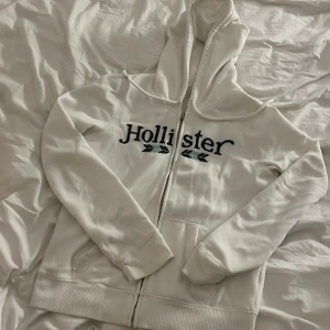 Vit hoodie från Hollister - Säljer en supermjuk och skön vit hoodie från Hollister. Den har en dragkedja framtill och en stor luva för extra mysfaktor. Perfekt för kyliga dagar eller bara för att chilla hemma. Hollister-loggan är snyggt broderad på bröstet. Passar perfekt till jeans eller mjukisbyxor!