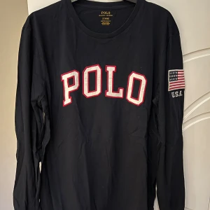 Blå långärmad t-shirt från Ralph Lauren - Säljer en blå långärmad T-shirt  från Ralph Lauren med 'POLO' tryckt i vitt och rött på framsidan. På ärmen finns en amerikansk flagga och texten 'U.S.A.'. Perfekt för en casual look eller en chill dag ute. Passar bra till jeans eller chinos! Storlek Medium. 