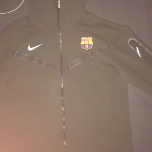 Svart Nike jacka med FC Barcelona-logga - Säljer en svart Nike jacka med FC Barcelona-logga på bröstet. Jackan har dragkedja och huva, perfekt för fotbollsfans. Den är långärmad och i bra skick. Passar både till vardags och träning.