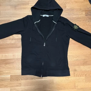 Stone island zip - Säljer en stilren svart zip från Stone Island med dragkedja och huva. Tröjan har en ikonisk logga på ärmen och praktiska fickor framtill. Perfekt för en avslappnad stil. Kontakta gärna vid frågor!