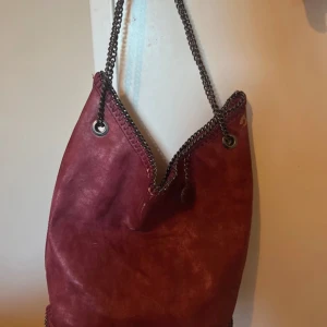 Röd axelväska från Stella McCartney - Snygg röd axelväska från Stella McCartney med kedjedetaljer runt kanterna och axelremmen. Väskan har en rund metallbricka med logotyp. Perfekt för att ge en stilren touch till din outfit. Inte 100% säker på att den är äkta då jag köpt den secound hand❤️lite sliten på viss ställen, se bild🥰hör av dig vid frågor