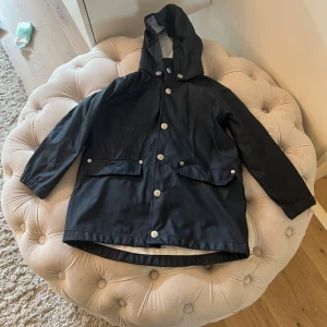 Svart regnjacka med huva - Säljer en svart regnjacka med huva och silverfärgade knappar. Jackan har långa ärmar och två stora fickor framtill. Perfekt för regniga dagar!