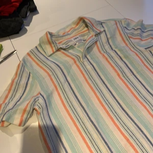 Randig skjorta från Zara - Säljer en färgglad randig skjorta från Zara i storlek M Skjortan har en avslappnad passform med korta ärmar och knappar framtill. Perfekt för en somrig look med sina ränder i blått, orange, grönt och vitt.