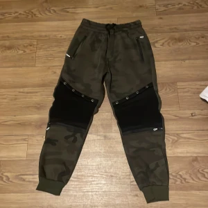 Camouflage mjukisbyxor med dragkedja - Snygga mjukisbyxor i camouflage-mönster med praktiska dragkedjor på fickorna. Byxorna har en elastisk midja med snörning och ribbade muddar vid bensluten. Perfekta för en avslappnad stil.