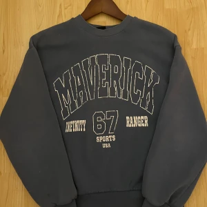 Blå sweatshirt från Gina Tricot - Säljer en blå sweatshirt från Gina Tricot med trycket 'Maverick Infinity 67 Ranger Sports USA' i vitt. Tröjan har långa ärmar och en rund halsringning. Perfekt för en avslappnad stil.