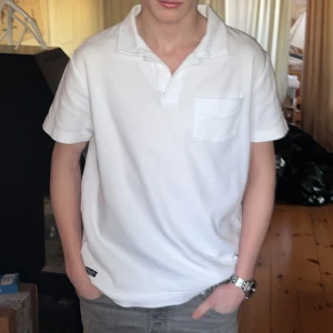 Vit kortärmad t-shirt från Robbie Moor - Säljer en stilren vit t-shirt från Robbie Moor med korta ärmar och en klassisk krage. T-shirten har en liten bröstficka och är perfekt för en avslappnad look. Märkesdetalj vid nederkanten ger en extra touch. 100% Bomull                                        Storlek: small S Bröst: 50cm Ärm: 5cm Längd: 70cm