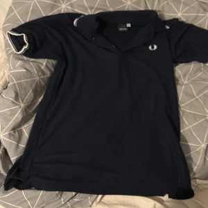 Mörkblå pikétröja från Fred Perry  - Säljer en klassisk mörkblå pikétröja från Fred Perry med vita detaljer på kragen och ärmsluten. Tröjan har korta ärmar och den ikoniska loggan broderad på bröstet. Perfekt för en stilren och avslappnad look.