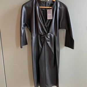 Svart klänning från Hermosa - Snygg svart klänning i syntetmaterial från Hermosa. Mycket smickrande 🤩🎉❤️Den har en omlott-design med en knutdetalj framtill och trekvartsärmar. Perfekt för en stilren look.