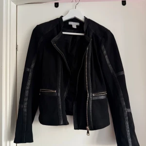 Svart jacka från H&M - Snygg svart jacka från H&M med dragkedjor framtill och på ärmarna. Jackan har en cool design med detaljer i syntetmaterial och en figurnära passform. Perfekt för att ge din outfit en edgy touch.
