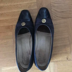 Svarta loafers från Margaux - Snygga svarta loafers från Margaux med en elegant gulddetalj på ovansidan. Skorna har en klassisk design med en låg klack och rundad tå, perfekt för en stilren look.