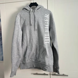 Grå hoodie från Tommy Hilfiger - Säljer en stilren grå hoodie från Tommy Hilfiger i ekologisk bomull. Den har en stor logga tryckt vertikalt på framsidan och en klassisk känguruficka. Perfekt för en avslappnad stil.
