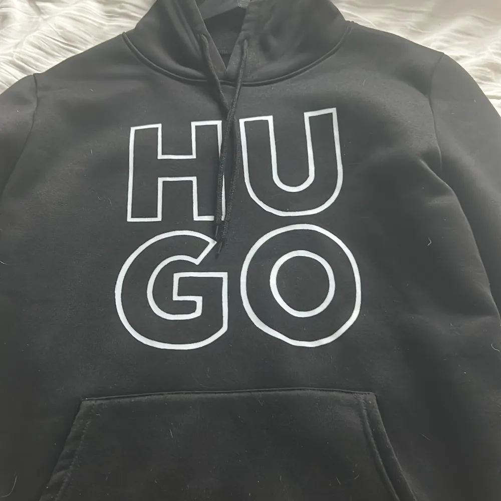 Säljer en stilren svart hoodie från Hugo med stor vit logga på framsidan. Tröjan har en klassisk känguruficka och justerbar huva med dragsko. Perfekt för en avslappnad look.. Neuletakit & Villapaidat.