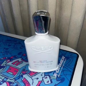 Creed Silver Mountain Water Parfym - Säljer en exklusiv Creed Silver Mountain Water parfym i en elegant vit flaska med silverdetaljer. Flaskan rymmer 100 ml och har en stilren design. Doften är känd för sina friska och rena toner, inspirerad av bergens friska luft och klara vatten. Knappt använd, dock lite trasig där man spruta som syns på bilden men funkar fortfarande perfekt 