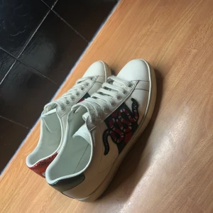 Vita sneakers med ormmotiv från Gucci - Snygga vita sneakers från Gucci med ett coolt ormmotiv på sidan. Skorna har klassiska snören och detaljer i rött och grönt på hälen. Har gått sönder lite fram vid sömmen . Bilderna på annonsen är lite gammla ( kan skicka bild hur det ser ut just nu)