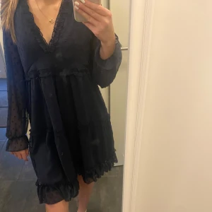 Mörkblå klänning med volangdetaljer - Snygg mörkblå klänning med v-ringning och volangdetaljer. Klänningen har långa ärmar och ett prickigt mönster som ger en elegant touch. Perfekt för både vardag och festliga tillfällen.