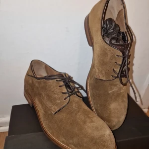 Bruna mockaskor från Massimo Dutti - Snygga bruna mockaskor från Massimo Dutti med klassisk snörning. Skorna har en stilren design med rund tå och en slitstark sula. Perfekta för en elegant look. Använd vid 1 tillfälle.