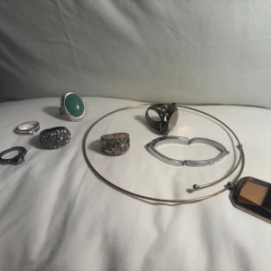 Samling av ringar och halsband - [KAN KÖPAS SEPARAT] 1 STERLING SILVERRING - 1 MINDRE SVART RING - 4 STÖRRE VINTAGERINGAR - 1 CHOKERHALSBAND MED STOR STEN. En elegant samling av smycken inklusive flera ringar och ett halsband. Ringarna har olika stilar, inklusive en med en stor grön sten och en med glittriga detaljer. Halsbandet har en minimalistisk design med en geometrisk hängande detalj.