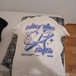 Vit t-shirt med lilla tryck - Säljer en vit t-shirt med ett coolt lilla tryck på både fram- och baksidan. T-shirten har korta ärmar och är i normal passform. Perfekt för en avslappnad stil.