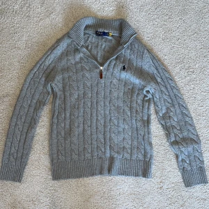 Grå halfzip tröja från Polo Ralph Lauren - Säljer en stilren grå stickad halfzip tröja från Polo Ralph Lauren med dragkedja och ribbade detaljer. Tröjan har ett klassiskt kabelstickat mönster och en broderad logotyp på bröstet. Perfekt för kyligare dagar. Använd endast 1 gång tidigare