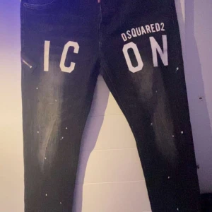 Svarta jeansbyxor från Dsquared2 - Snygga svarta jeansbyxor från Dsquared2 med texten 'ICON' tryckt på framsidan. Byxorna har en modern look med slitna detaljer och små vita färgstänk. Perfekta för en trendig stil.