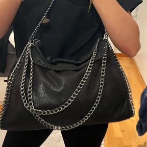 Svart axelväska med kedjedetaljer - Stor svart väska med ”glittrig” yta och kedjedetaljer. Väskan har en dragkedja för säker förvaring och en rymlig insida med olika fack. (Två små öppna, ett större öppet, och två lite större stängbara). Väldigt bra väska som är rymlig! Det enda som finns defekt på är en liten snörning typ som lossnat lite. 