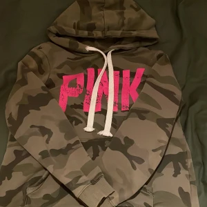 Camouflage hoodie med rosa tryck - Säljer en cool camouflage hoodie från Pink med ett stort rosa tryck på framsidan. Hoodien har en justerbar huva med vita snören och en klassisk känguruficka. Perfekt för en avslappnad stil.