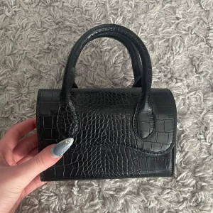 Svart liten handväska med krokodilmönster - Snygg svart liten handväska med krokodilmönster. Den har två handtag och en stilren design som passar perfekt för en chic look. Väskan är kompakt men rymmer det viktigaste för en dag ute.