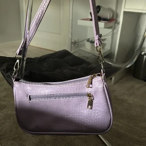 Ljuslila axelväska med krokodilmönster - Snygg ljuslila axelväska med krokodilmönster och guldfärgade detaljer. Väskan har en dragkedja på framsidan. Perfekt för att ge din outfit en trendig touch.❤️