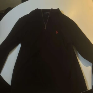 Svart tröja från Polo Ralph Lauren - Säljer en stilren svart tröja från Polo Ralph Lauren med dragkedja vid halsen och det klassiska logotypbroderiet i rött på bröstet. Perfekt för en avslappnad men ändå elegant look.