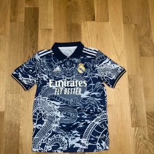 Real Madrid T-shirt - Säljer en supercool blå Real Madrid fotbollströja från Adidas med ett unikt drakmönster i vitt. Tröjan har korta ärmar och en klassisk krage. Perfekt för alla fans som vill sticka ut på läktaren eller planen! 🐉⚽köpte den för en vecka seda, har bara haft på mig den en gång, gick inte att returnera, behöver pengarna tillbaka därför säljer jag den, pris går att diskutera vid snabb affär