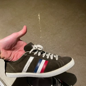 Bruna sneakers från Moncler - Snygga bruna sneakers från Moncler med vita snören och en klassisk design. Skorna har Monclers ikoniska ränder i rött, vitt och blått på sidan. Perfekta för en stilren look. Ganska använda där av priset storlek 44. Kvitto finns ej kvar med dust bags och kartong följer med. Kan gå ner lite i pris vid snabb och smidig affär!