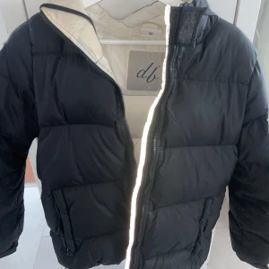 Svart dunjacka från db - Säljer en svart dunjacka från db med broderad logga på ärmen. Jackan har en dragkedja framtill och är långärmad. Perfekt för kyliga dagar med sin vadderade design. 
