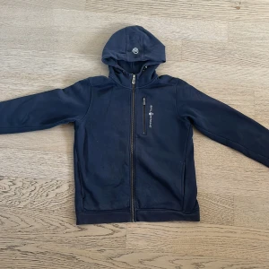 Mörkblå huvtröja från Sail Racing - Säljer en mörkblå huvtröja från Sail Racing med dragkedja framtill. Tröjan har en praktisk huva och en bröstficka med dragkedja. 