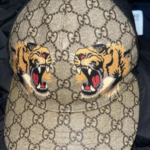 Mönstrad keps från Gucci - Snygg keps från Gucci med ett djärvt mönster av tigrar och det klassiska Gucci-mönstret. Kepsen har en justerbar rem baktill för perfekt passform.