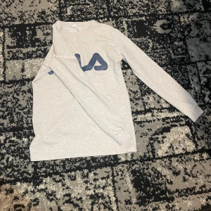 Grå långärmad tröja från FILA - Säljer en stilren grå långärmad tröja från FILA med deras ikoniska logga i blått och rött på bröstet. Perfekt för en avslappnad look.