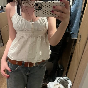 Vit Brandy Melville topp med knappar - Säljer en söt vit brandy melville topp med knappar framtill. Den har en luftig passform och justerbara axelband! Perfekt för sommar/vår eller med kofta. Passar s-m