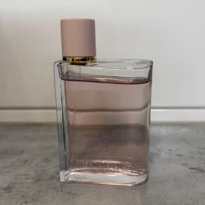 Parfym från Burberry - Elegant parfym från Burberry i en stilren, rektangulär flaska med en ljusrosa ton. Flaskan har en mjuk rosa kork med en gyllene ringdetalj. Perfekt för den som älskar en sofistikerad och modern doft.