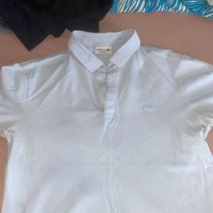 Vit pikétröja från Lacoste - Säljer en klassisk vit pikétröja från Lacoste i regular fit. Tröjan har korta ärmar och en broderad logotyp på bröstet. Perfekt för en stilren och avslappnad look.