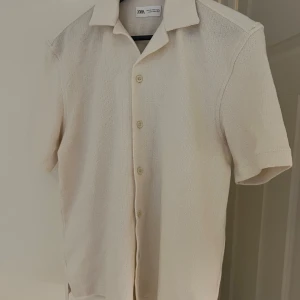 Beige kortärmad skjorta från Zara - Snygg beige skjorta från Zara med korta ärmar och knappar framtill. Perfekt för en stilren och avslappnad look. Skjortan har en klassisk krage och är tillverkad i ett bekvämt material. Fick den som present den är för stor på mig, perfekt nu för våren och sommaren!