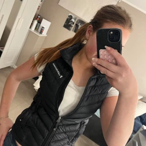 Svart dunväst från Peak Performance - Snygg svart dunväst från Peak Performance med dragkedja framtill. Perfekt för lager-på-lager och håller dig varm under kyliga dagar. Västen har en stilren design med diskret logga på bröstet.