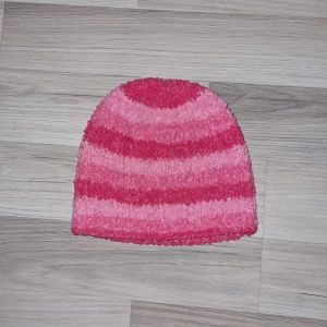 Rosa randig mössa - Säljer en mysig rosa mössa med ränder i olika nyanser av rosa. Perfekt för att hålla värmen under kyliga dagar. Den är mjuk och bekväm att bära. Mössan är hemmastickade.