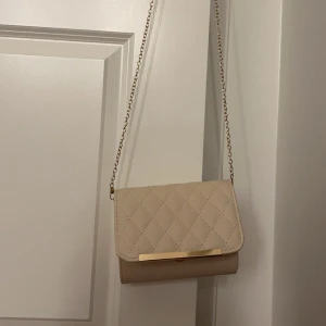 Beige quiltad axelväska med kedja - Snygg beige axelväska med quiltat mönster och guldfärgad kedja. Har aldrig använts. Kedjan är justerbar. 